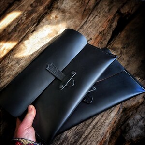 May include: Two black leather document holders with flap closures and straps secured by metal rivets. The holders are displayed on a rustic wooden surface. The leather has a smooth finish and visible stitching.
