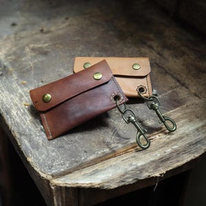May include: Two small leather wallets in shades of brown and tan, each with a snap closure and a metal clasp. The wallets are displayed on a weathered wooden surface, showcasing their handcrafted details and vintage aesthetic.