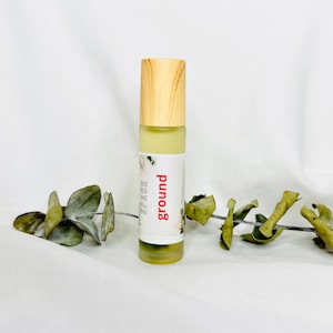 Puede incluir: Un frasco de aceite esencial roll-on con tapa de madera clara y una etiqueta blanca con la palabra "ground" en rojo. El frasco contiene un líquido verde claro. El fondo es blanco con una ramita de eucalipto seco.