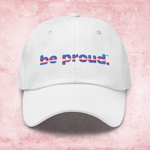 May include: White baseball cap with the text "be proud." in blue letters with a transgender pride flag design.