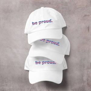 May include: Three white baseball caps stacked on top of each other. Each cap has the phrase "be proud." embroidered in blue, pink, and white.