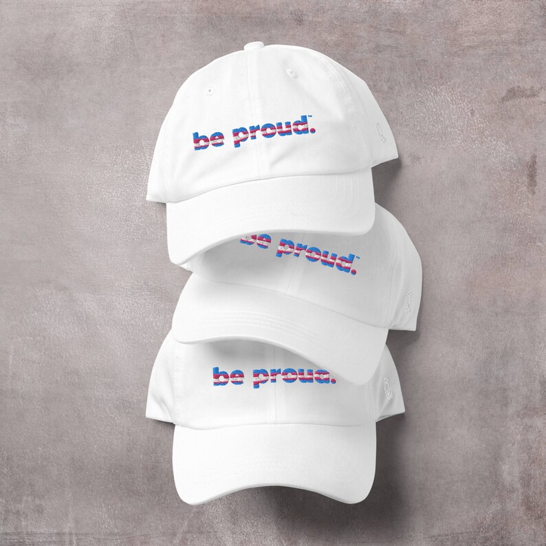 May include: Three white baseball caps stacked on top of each other. Each cap has the phrase "be proud." embroidered in blue, pink, and white.
