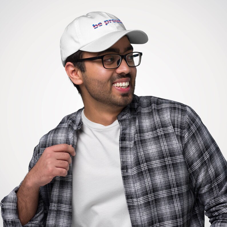 May include: A person wearing a white baseball cap with the text "be proud" in blue and red letters. The person is wearing a white t-shirt and a black and white plaid shirt.