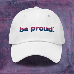 May include: White baseball cap with the text "be proud." in blue, pink, and purple lettering.