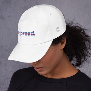 May include: White baseball cap with the text "Be proud." in blue, red, and white lettering. The text is in a stylized font with a thin blue line above and below the text.