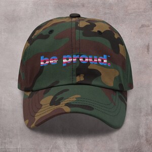 May include: A camouflage baseball cap with the text "be proud." embroidered in blue, pink, and white.