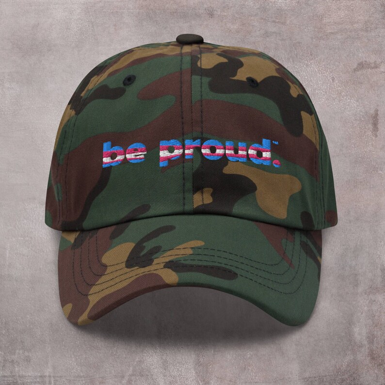 May include: A camouflage baseball cap with the text "be proud." embroidered in blue, pink, and white.