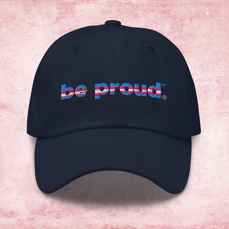 May include: A navy blue baseball cap with the text "be proud." embroidered in white, pink, and blue. The text is stylized to look like the transgender pride flag.