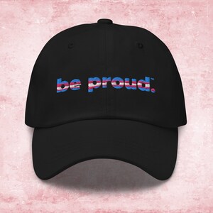 May include: Black baseball cap with the text "be proud." in blue and pink letters, with a transgender pride flag design.