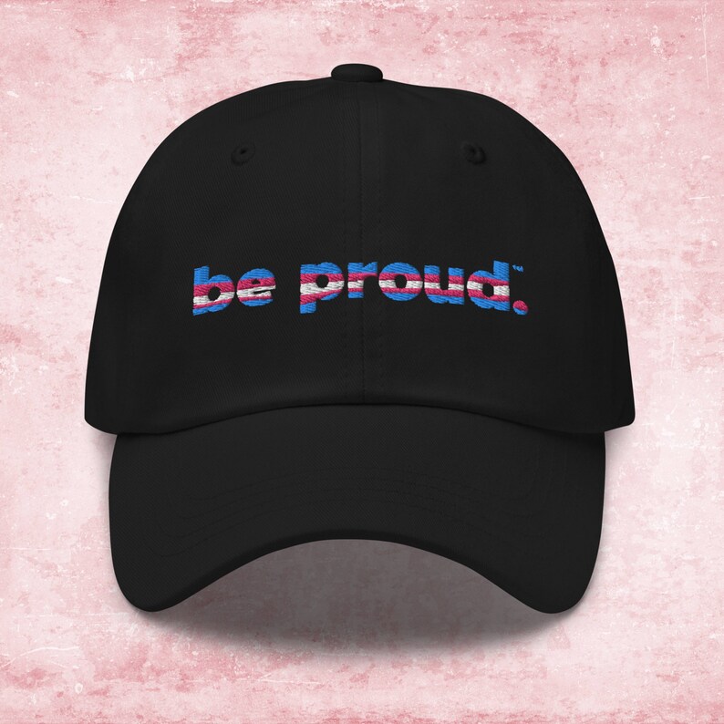 May include: Black baseball cap with the text "be proud." in blue and pink letters, with a transgender pride flag design.