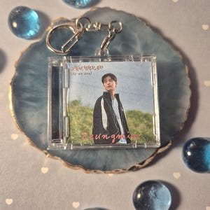 Könnte beinhalten: Ein durchsichtiger Kunststoff-Schlüsselanhänger mit einem kleinen CD-Hüllen-Einsatz. Der Einsatz zeigt eine Person in einer schwarz-weißen Jacke. Der Name "Seungmin" ist in rosa Schrift geschrieben. Der Schlüsselanhänger hat einen silbernen Metallverschluss.