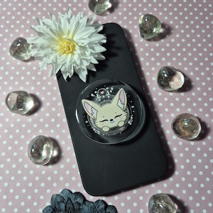 May include: A black mobile phone with a clear pop socket showing a cartoon fox face. The pop socket has a white and pink design. The background is pink with white polka dots, and there are clear, rounded stones and a white flower.