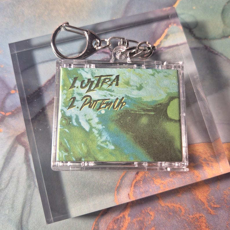 Peut inclure: Porte-cl&eacute;s en plastique transparent avec un anneau et une cha&icirc;ne en m&eacute;tal argent&eacute;. Le porte-cl&eacute;s pr&eacute;sente un motif abstrait vert et bleu avec le texte "1. ULTRA" et "2. PUTTEM UP" en lettres dor&eacute;es.