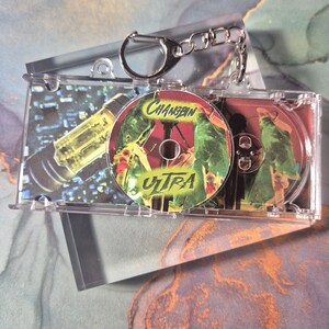 Peut inclure: Un porte-cl&eacute;s en plastique transparent avec un mini &eacute;tui &agrave; CD. Le CD &agrave; l'int&eacute;rieur pr&eacute;sente un design vert et rouge avec le texte "Changbin ULTRA". L'&eacute;tui est ouvert, r&eacute;v&eacute;lant le CD et un insert imprim&eacute; avec un design jaune et noir.