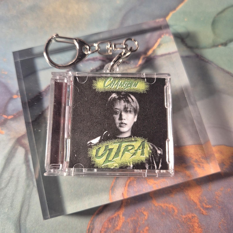 Peut inclure: Un porte-cl&eacute;s en plastique transparent avec un petit bo&icirc;tier de CD contenant une photo d'une personne et le mot "ULTRA" en vert. Le porte-cl&eacute;s a un fermoir et une cha&icirc;ne en m&eacute;tal argent&eacute;.