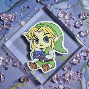 May include: A sticker featuring a cartoon character with blonde hair, a green hat, and green tunic, holding a blue video game controller. The character has large blue eyes and pointed ears. The sticker is on a clear, square surface.