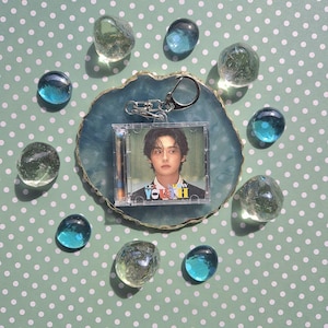 Puede incluir: Un llavero de plástico transparente con una tarjeta de foto del tamaño de un CD que presenta a un joven con cabello oscuro. La tarjeta de foto tiene el texto "Lee Know" y "Youth" en letras de colores. El llavero está sobre un fondo de lunares blancos y verdes.