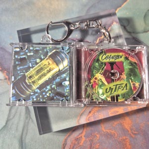 Peut inclure: Un porte-cl&eacute;s en plastique transparent avec un mini &eacute;tui &agrave; CD. L'&eacute;tui contient un CD avec le texte "Changbin Ultra" en vert et jaune. L'autre c&ocirc;t&eacute; de l'&eacute;tui montre un objet cylindrique jaune.