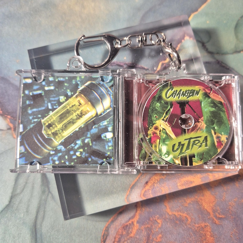 Peut inclure: Un porte-cl&eacute;s en plastique transparent avec un mini &eacute;tui &agrave; CD. L'&eacute;tui contient un CD avec le texte "Changbin Ultra" en vert et jaune. L'autre c&ocirc;t&eacute; de l'&eacute;tui montre un objet cylindrique jaune.