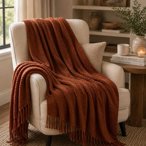 May include: A rust-colored knitted throw blanket with a diagonal ribbed texture and fringe detailing along the edges. The blanket is draped over a cream-colored upholstered armchair, showcasing its soft fabric and long, hanging tassels.