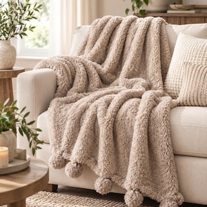 May include: A beige faux fur throw blanket with a thick, shaggy texture draped over a sofa. The blanket features a series of matching beige pom-poms attached along the bottom edge, creating a decorative border with a soft, plush appearance.