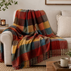 Può includere: Una coperta in tessuto con un motivo a quadri grandi nei toni del rosso intenso, giallo senape, blu scuro e marrone. Il tessuto ha una superficie ruvida e testurizzata, rifinita con lunghe frange annodate lungo il bordo inferiore.