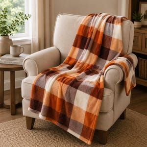 May include: A plush throw blanket featuring a large-scale plaid pattern in shades of burnt orange, dark brown, and cream. The soft, fuzzy fabric is draped over a light beige upholstered armchair, showcasing the thick texture and checkered design.