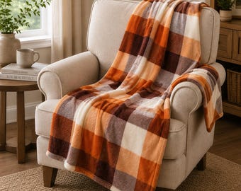 May include: A plush throw blanket featuring a large-scale plaid pattern in shades of burnt orange, dark brown, and cream. The soft, fuzzy fabric is draped over a light beige upholstered armchair, showcasing the thick texture and checkered design.