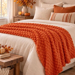 May include: A vibrant orange chunky knit throw blanket with a textured pom-pom pattern draped over a bed with cream-coloured bedding. The blanket features a thick, soft yarn construction with a repeating spherical design across the entire surface.