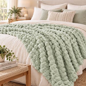 May include: A sage green chunky knit throw blanket with a textured pom-pom design draped over a bed. The blanket features a thick, soft yarn construction with rows of spherical tufts, providing a dense and tactile surface across the bedding.