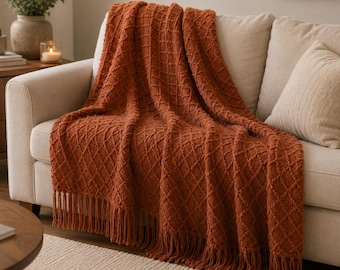 May include: A burnt orange throw blanket featuring a raised diamond texture pattern and long fringe trim along the bottom edge. The blanket is draped over a light beige sofa, showcasing the intricate knit design and the deep, warm color.