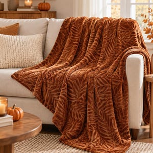 Può includere: Una coperta in peluche color arancio bruciato drappeggiata su un divano di colore chiaro. La coperta presenta un motivo testurizzato e in rilievo di foglie simili a felci che si ripetono su tutta la superficie, creando un aspetto morbido e vellutato.