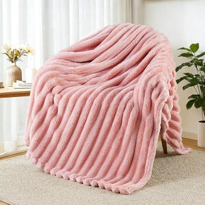 May include: A soft, plush, light pink throw blanket with a textured, ribbed, or channel-tufted pattern. The blanket is draped over a chair, showcasing its thick, fuzzy material and consistent vertical lines that run across the entire surface.