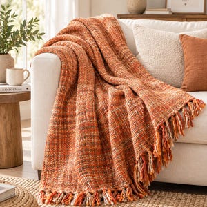 May include: A chunky knit throw blanket in shades of burnt orange, rust, and cream with a textured weave. The blanket features long, twisted fringe trim along the bottom edge and is draped over the arm of a white sofa with a soft, thick fabric.