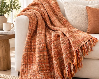 May include: A chunky knit throw blanket in shades of burnt orange, rust, and cream with a textured weave. The blanket features long, twisted fringe trim along the bottom edge and is draped over the arm of a white sofa with a soft, thick fabric.