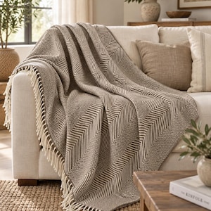 May include: A woven throw blanket featuring a repeating pattern of taupe and white chevron stripes and small geometric squares. The blanket has a textured surface and is finished with long, cream-colored fringe along the bottom edge.