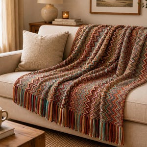 Può includere: Una coperta in maglia rettangolare caratterizzata da un motivo a chevron multicolore nelle tonalità ruggine, ottanio, crema e marrone. La coperta presenta una superficie testurizzata ed è rifinita con lunghe frange multicolore sul bordo inferiore.