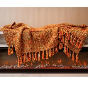 May include: A woven throw blanket in shades of orange, yellow, and green with long fringe. The blanket is draped over a white shelf above a fireplace with a glowing fire effect.