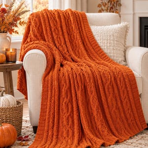 May include: A vibrant orange knitted throw blanket featuring a prominent vertical cable knit pattern and a ribbed border. The blanket is draped over a light-colored upholstered armchair, showcasing its thick, textured yarn construction.