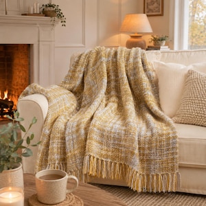 May include: A woven throw blanket featuring a plaid pattern in shades of mustard yellow, white, and grey. The blanket has a textured, chunky knit appearance with long, knotted fringe detailing along the bottom edge, draped over a light-colored sofa.
