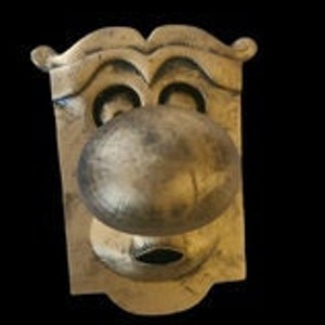 May include: A vintage-style brass door knob with a face design. The knob has a large, round center and a rectangular backplate with a stylized mustache and eye shapes. The finish is aged and tarnished.