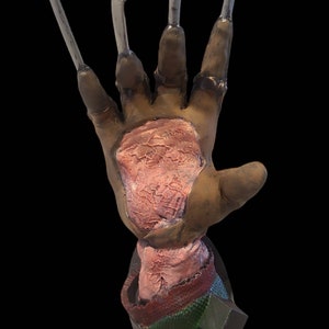 May include: A prop hand with metal claws, featuring a brown and pink color scheme. The hand has a textured appearance, with visible details on the skin and the base of the claws. The base of the hand has a green and red striped pattern.