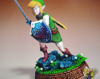 May include: A detailed figurine of a character from The Legend of Zelda. The figure is posed with a sword and shield, wearing a green tunic, hat, and brown boots. The base is decorated with a detailed pattern and a blue and green landscape.