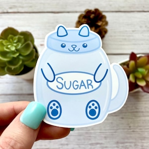May include: A light blue sticker in the shape of a cat-like sugar container. The sticker has the word "SUGAR" in a white oval. The cat-shaped container has ears, paws, and a tail. The background is a light wood surface with plants.