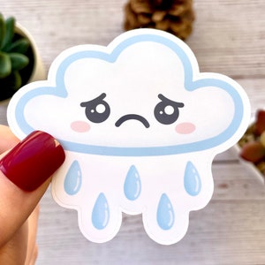 May include: A white cloud sticker with a sad face, light blue outline, and light blue raindrops. The cloud has pink cheeks and a curved black mouth. The sticker is held by a person with red nail polish.
