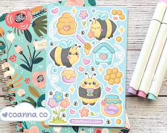 May include: A sheet of bee-themed stickers with various designs, including bees, honeycombs, and honey pots, on a light blue background. The stickers are surrounded by small hearts, flowers, and stars. The stickers are placed on top of a floral notebook.