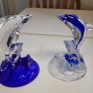 Peut inclure: Deux figurines de dauphins en verre dans des poses bondissantes. Un dauphin est en verre transparent sur une base en verre bleu, tandis que l'autre est en verre transparent avec une bande bleue sur le dos, sur une base en verre transparent.