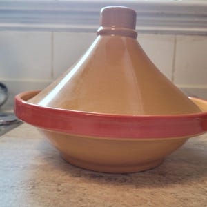 Op de afbeelding: Een keramische tajine met een conisch deksel en een brede basis. Het hoofdgedeelte en het deksel zijn afgewerkt met een gladde, glanzende beige glazuurlaag, terwijl de buitenrand van de basis is geaccentueerd met een levendige, rode band.