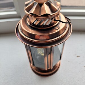 May include: A copper-coloured metal lantern featuring a circular top with a fluted design, a vented chimney section, and a glass-panelled body. The lantern includes a thin black wire handle attached to the side and a circular base.