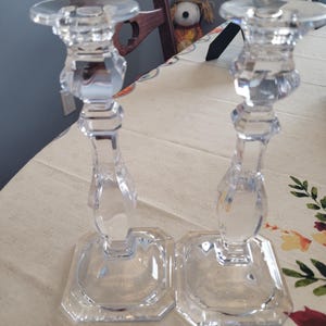 May include: A pair of clear glass candle holders featuring a faceted, multi-sided stem design and a wide, octagonal base. Each holder has a circular top platform for a candle, with a transparent, reflective finish and a structured, geometric shape.